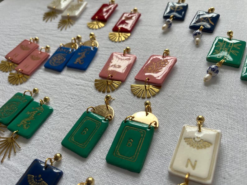 May include: A collection of colorful earrings with gold-colored accents. The earrings feature rectangular and square shapes in various colors, including red, pink, green, and blue. Some earrings have gold-colored fan-shaped details and decorative patterns.