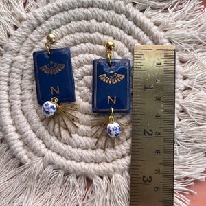May include: A pair of handmade earrings. Each earring features a dark blue rectangular pendant with gold accents and the letter "N". Below the pendant is a gold-colored starburst design with a small blue and white bead. The earrings are approximately 1.5 inches long.