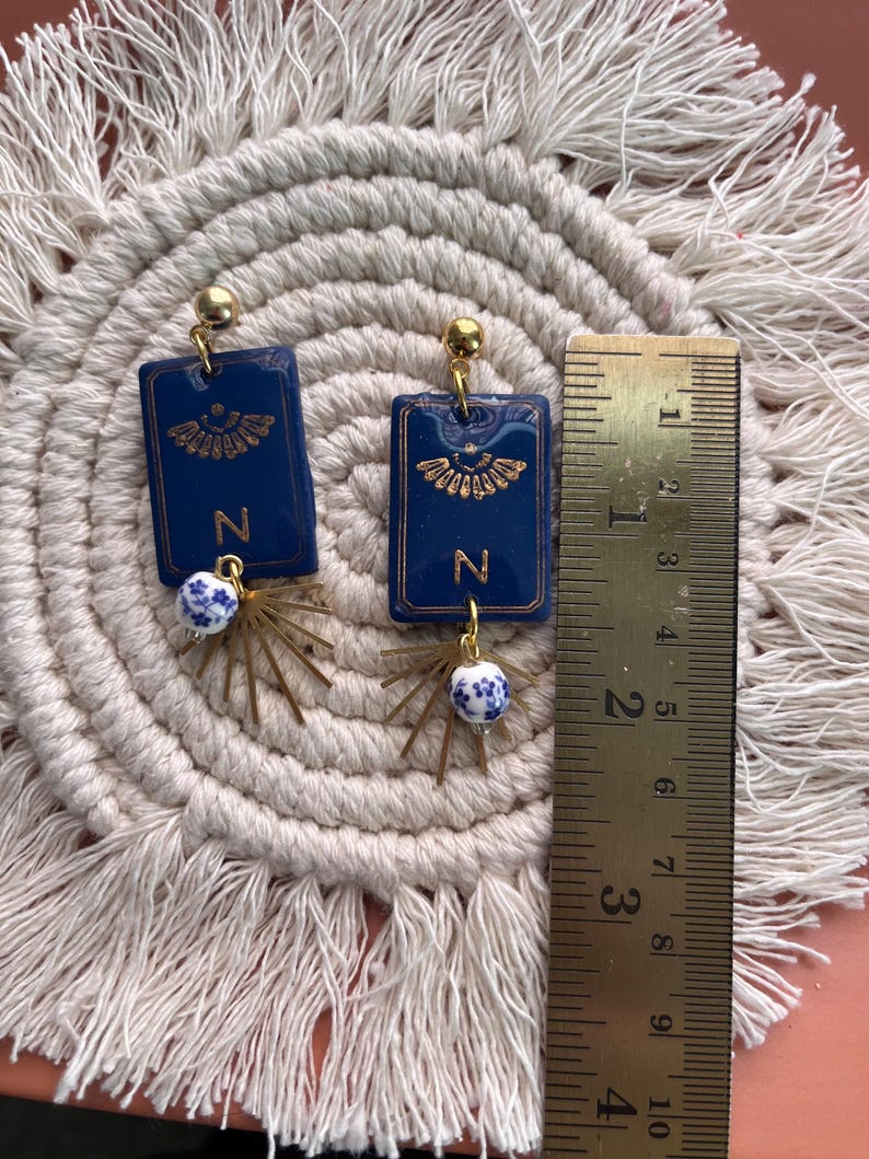 May include: A pair of handmade earrings. Each earring features a dark blue rectangular pendant with gold accents and the letter "N". Below the pendant is a gold-colored starburst design with a small blue and white bead. The earrings are approximately 1.5 inches long.