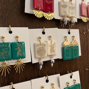 May include: A display of handmade earrings with various designs. The earrings feature rectangular and fan-shaped elements in red, white, pink, and teal. Some earrings have gold accents and the letter "N" or the word "JOKER".