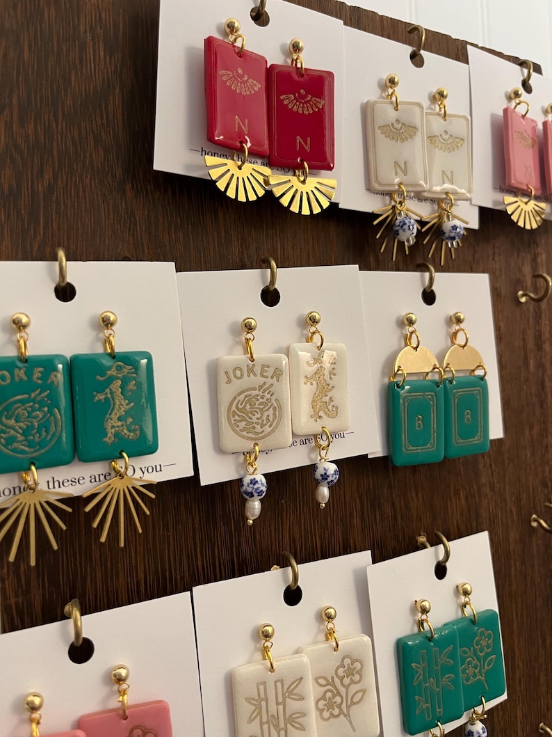 May include: A display of handmade earrings with various designs. The earrings feature rectangular and fan-shaped elements in red, white, pink, and teal. Some earrings have gold accents and the letter "N" or the word "JOKER".