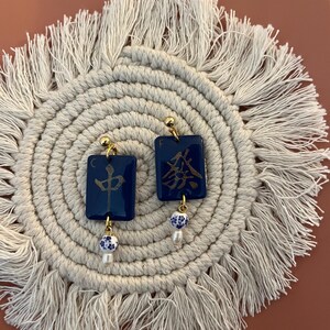 May include: A pair of dark blue square earrings with gold-colored accents. Each earring features gold Chinese characters and a small, dangling white and blue bead with a pearl. The earrings are displayed on a cream-colored, circular woven mat.