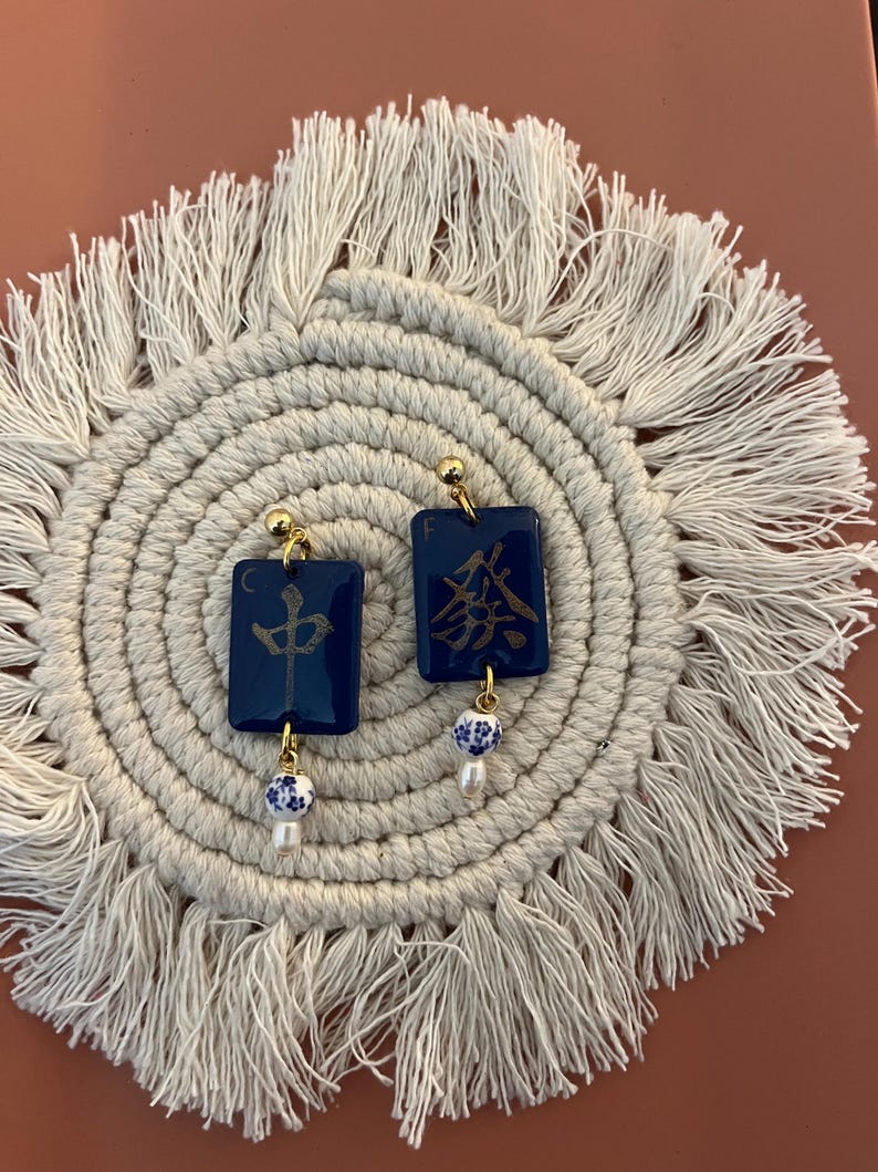 May include: A pair of dark blue square earrings with gold-colored accents. Each earring features gold Chinese characters and a small, dangling white and blue bead with a pearl. The earrings are displayed on a cream-colored, circular woven mat.