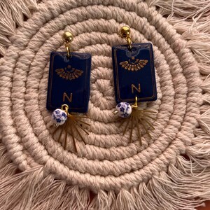 May include: A pair of navy blue rectangular earrings with gold accents. Each earring features a gold letter "N" and a decorative gold design. The earrings have a gold ball at the top and a white and blue bead with a gold starburst charm.