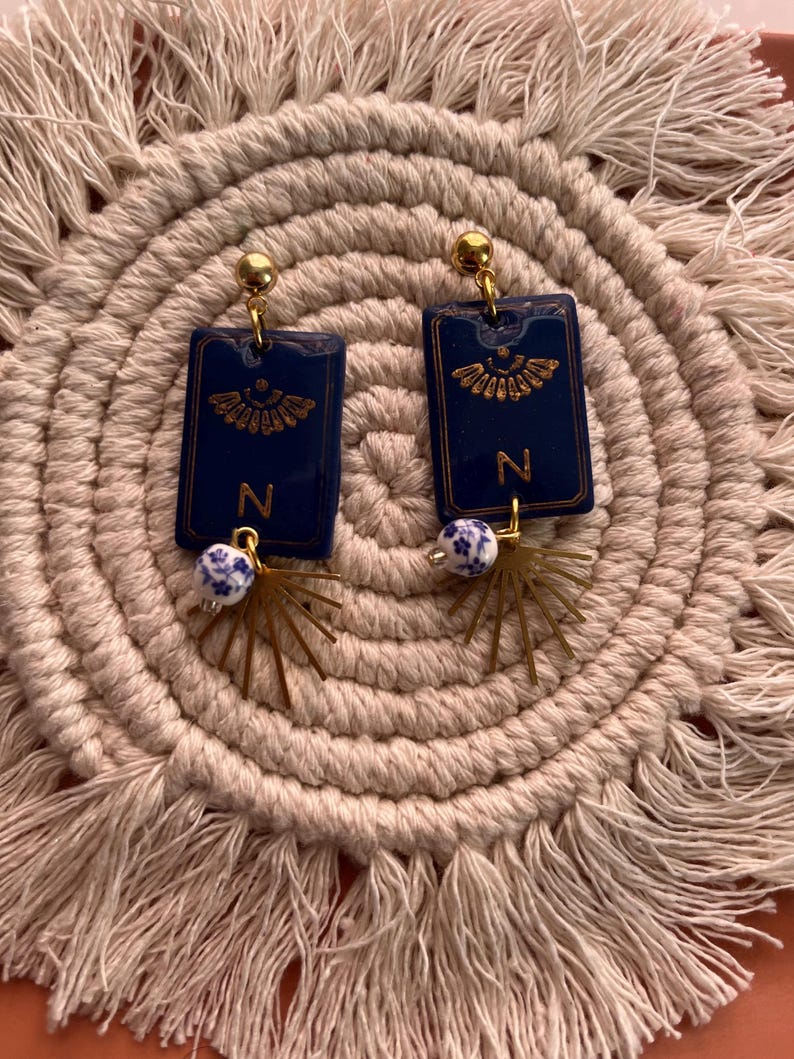 May include: A pair of navy blue rectangular earrings with gold accents. Each earring features a gold letter "N" and a decorative gold design. The earrings have a gold ball at the top and a white and blue bead with a gold starburst charm.