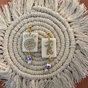 May include: A pair of white rectangular earrings with gold accents. One earring features the word "JOKER" and a gold design, while the other has a gold dragon design. Each earring has a blue and white bead and a small pearl. The earrings are displayed on a cream-colored, fringed mat.