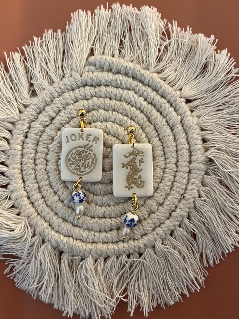 May include: A pair of white rectangular earrings with gold accents. One earring features the word "JOKER" and a gold design, while the other has a gold dragon design. Each earring has a blue and white bead and a small pearl. The earrings are displayed on a cream-colored, fringed mat.