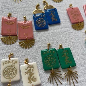 May include: A collection of colorful, handmade earrings. The earrings feature rectangular and fan-shaped elements in various colors, including pink, blue, green, and white. Some earrings have the word "JOKER" or the letter "N" printed on them, along with gold-colored dragon and sunburst designs.
