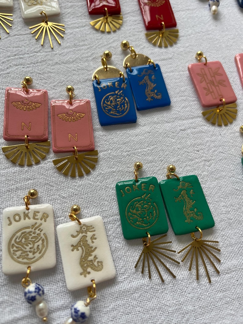 May include: A collection of colorful, handmade earrings. The earrings feature rectangular and fan-shaped elements in various colors, including pink, blue, green, and white. Some earrings have the word "JOKER" or the letter "N" printed on them, along with gold-colored dragon and sunburst designs.