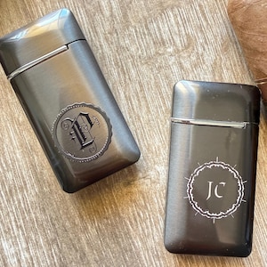 Puede incluir: Dos pitilleras de color gris plomo con dise&ntilde;os circulares decorativos. Una pitillera presenta la letra "P" y la otra las iniciales "JC". Las pitilleras son rectangulares con tapa abisagrada.