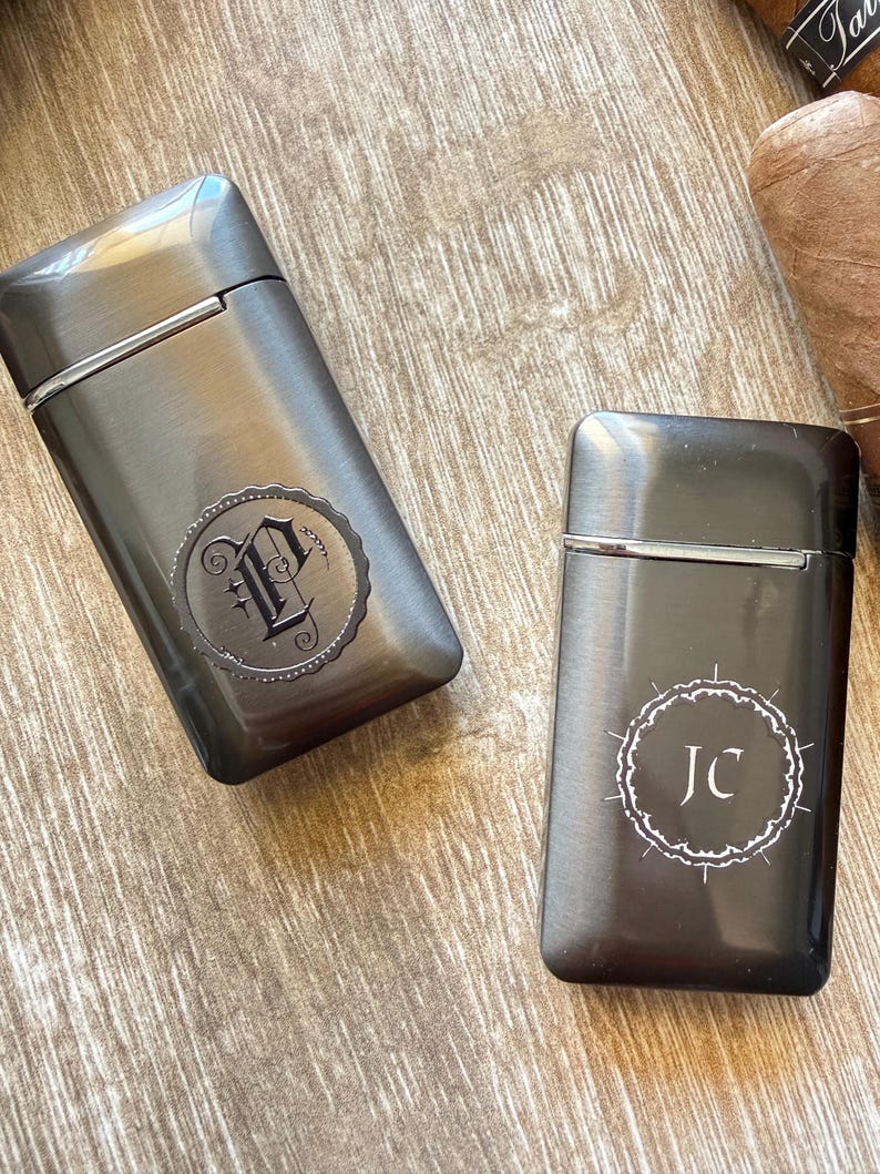 Puede incluir: Dos pitilleras de color gris plomo con dise&ntilde;os circulares decorativos. Una pitillera presenta la letra "P" y la otra las iniciales "JC". Las pitilleras son rectangulares con tapa abisagrada.