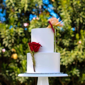 Puede incluir: Un pastel de boda blanco de dos niveles decorado con una sola rosa roja y una rosa melocotón en el lateral. El pastel está sobre un soporte de pastel blanco y una mesa blanca.