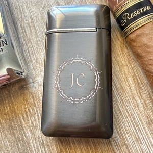 Puede incluir: Un encendedor de metal gris oscuro con las iniciales "JC" dentro de un borde decorativo. Un cigarro con una etiqueta marr&oacute;n y dorada es visible en el fondo. El encendedor tiene forma rectangular con bordes redondeados.