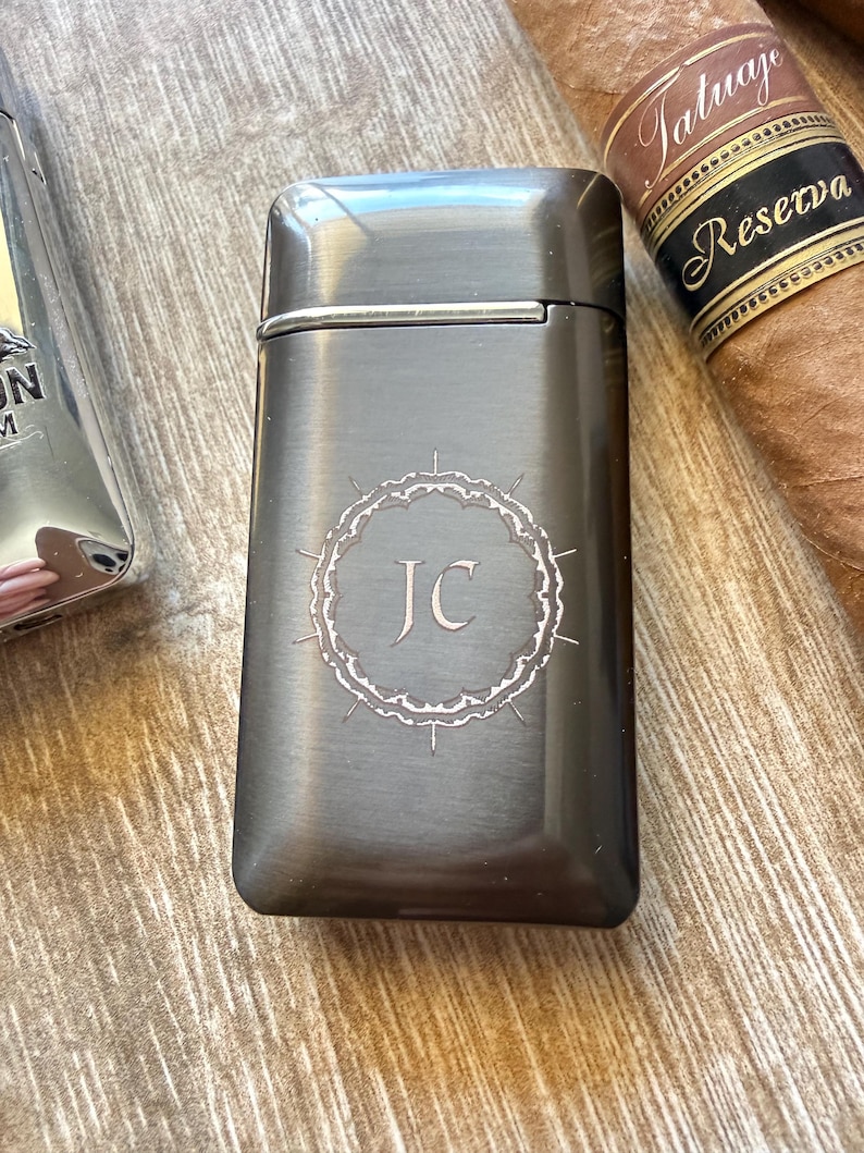 Puede incluir: Un encendedor de metal gris oscuro con las iniciales "JC" dentro de un borde decorativo. Un cigarro con una etiqueta marr&oacute;n y dorada es visible en el fondo. El encendedor tiene forma rectangular con bordes redondeados.
