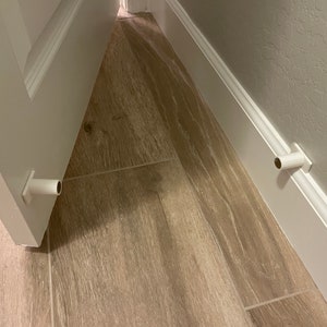 May include: Two white plastic door stops are installed on the floor, one on each side of a doorway. The door stops are rectangular with a rounded cylinder protruding from the top.