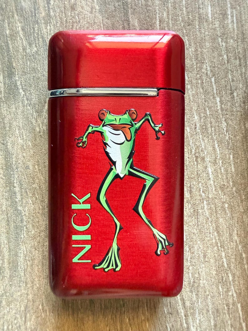 Puede incluir: Un encendedor de metal rojo con tapa abatible. El encendedor presenta una ilustraci&oacute;n de rana de dibujos animados verde y blanca con el nombre "NICK" impreso verticalmente en verde. El encendedor tiene un acabado de metal cepillado.