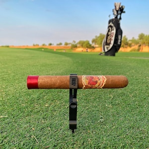 May include: A black cigar holder with the word "GRIP" printed on it, holding a brown cigar with a red band and a label. The cigar is on a green grassy surface with a golf bag in the background.