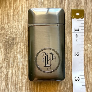 Puede incluir: Un encendedor rectangular de metal cepillado con un emblema decorativo con la letra "P" en una fuente adornada. El encendedor mide aproximadamente 7,6 cm de alto, con una cinta m&eacute;trica dorada y blanca al lado.
