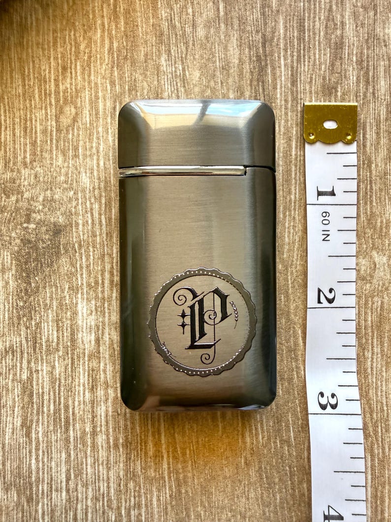 Puede incluir: Un encendedor rectangular de metal cepillado con un emblema decorativo con la letra "P" en una fuente adornada. El encendedor mide aproximadamente 7,6 cm de alto, con una cinta m&eacute;trica dorada y blanca al lado.