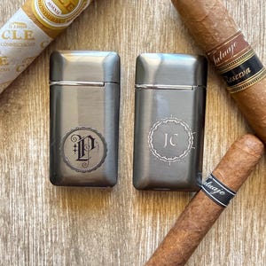 Puede incluir: Dos encendedores de metal cepillado con iniciales grabadas, uno con una "P" y el otro con "JC", se muestran con varios cigarros. Los cigarros tienen envoltorios marrones y bandas negras con texto blanco. Los encendedores son rectangulares.