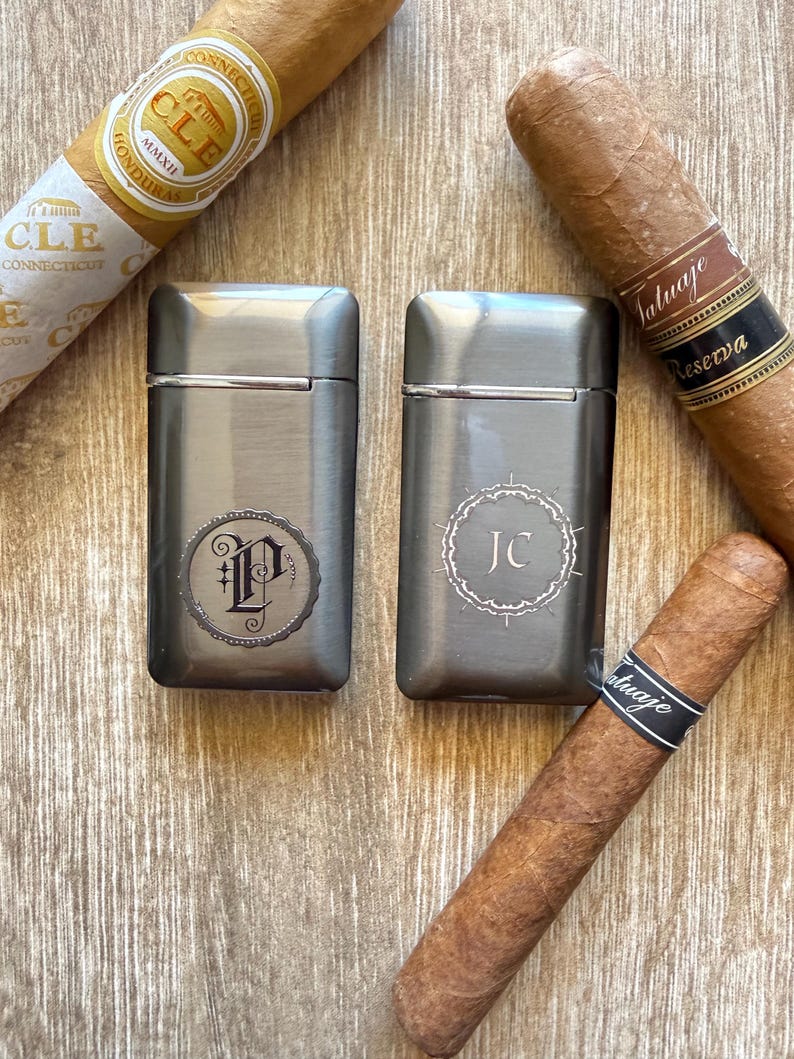 Puede incluir: Dos encendedores de metal cepillado con iniciales grabadas, uno con una "P" y el otro con "JC", se muestran con varios cigarros. Los cigarros tienen envoltorios marrones y bandas negras con texto blanco. Los encendedores son rectangulares.