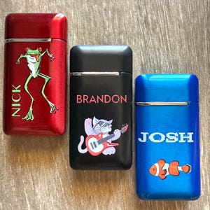 Puede incluir: Tres encendedores personalizados en rojo, negro y azul. El encendedor rojo presenta un gr&aacute;fico de rana verde y el nombre "NICK". El encendedor negro tiene un gato tocando la guitarra y el nombre "BRANDON". El encendedor azul muestra un pez payaso y el nombre "JOSH".