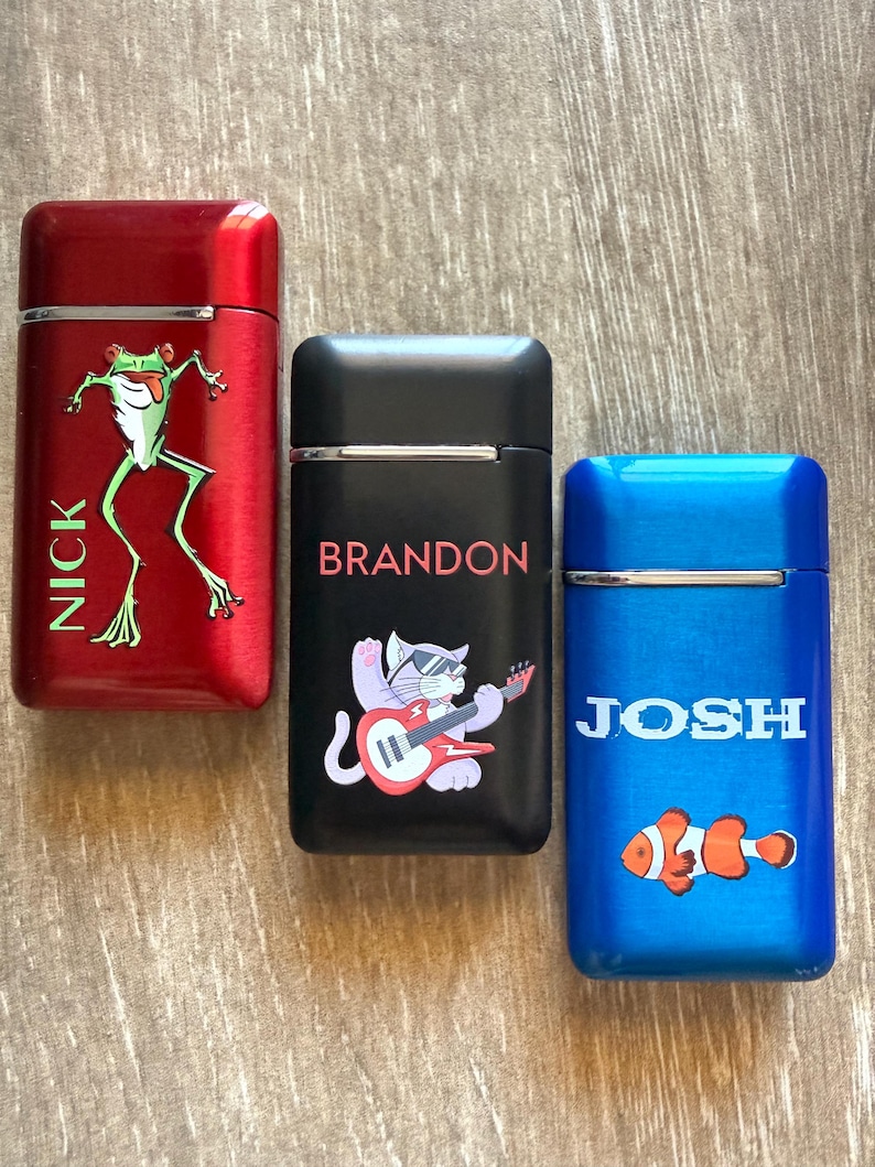 Puede incluir: Tres encendedores personalizados en rojo, negro y azul. El encendedor rojo presenta un gr&aacute;fico de rana verde y el nombre "NICK". El encendedor negro tiene un gato tocando la guitarra y el nombre "BRANDON". El encendedor azul muestra un pez payaso y el nombre "JOSH".