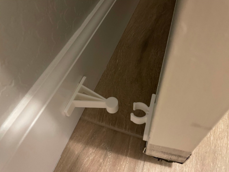 May include: White plastic door stop with a hook and a lever. The door stop is attached to a white door and the lever is attached to the floor.