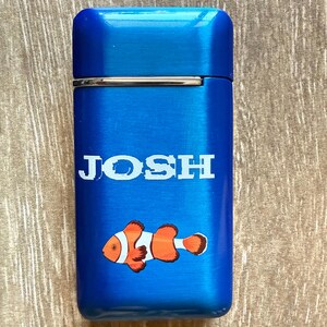 Puede incluir: Un encendedor azul con el nombre "JOSH" en letras blancas. Debajo del nombre hay un pez payaso de dibujos animados en naranja, blanco y negro. El encendedor tiene un acabado de metal cepillado.