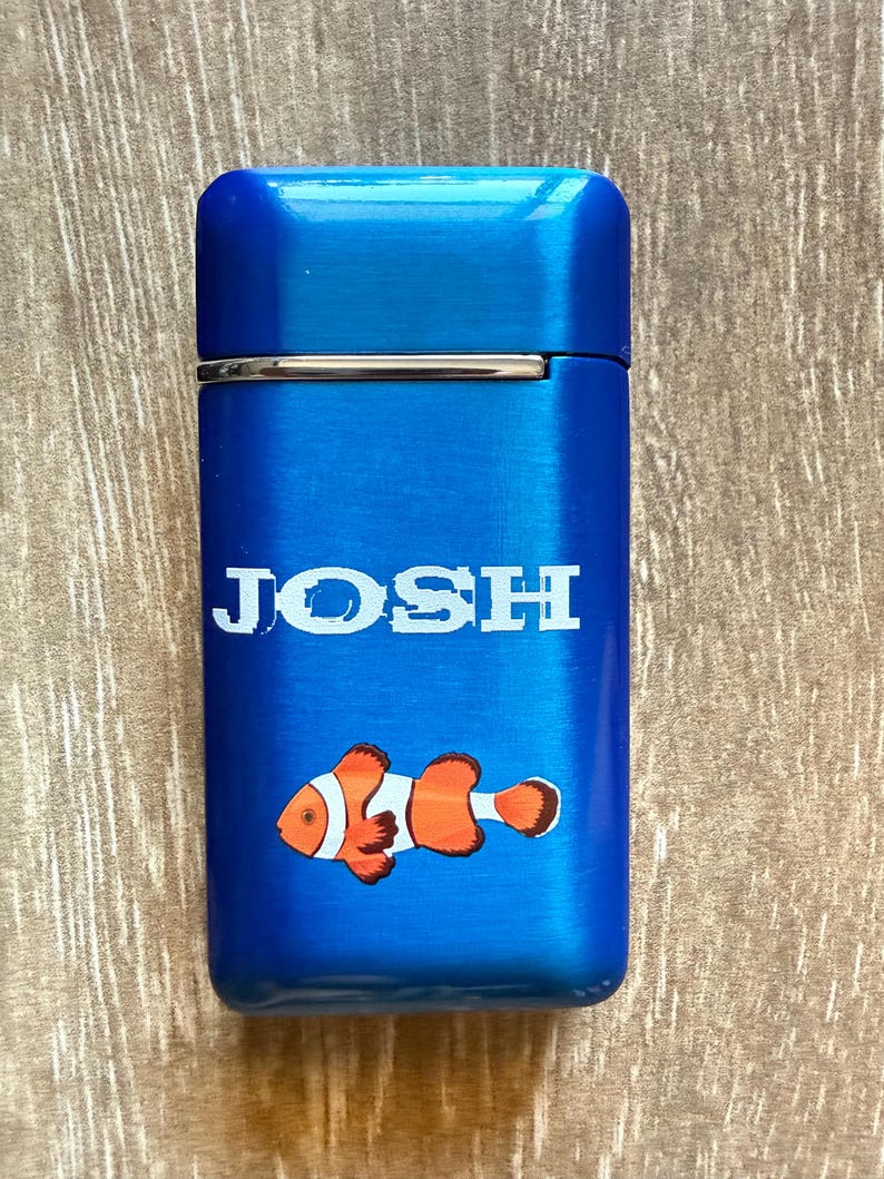 Puede incluir: Un encendedor azul con el nombre "JOSH" en letras blancas. Debajo del nombre hay un pez payaso de dibujos animados en naranja, blanco y negro. El encendedor tiene un acabado de metal cepillado.