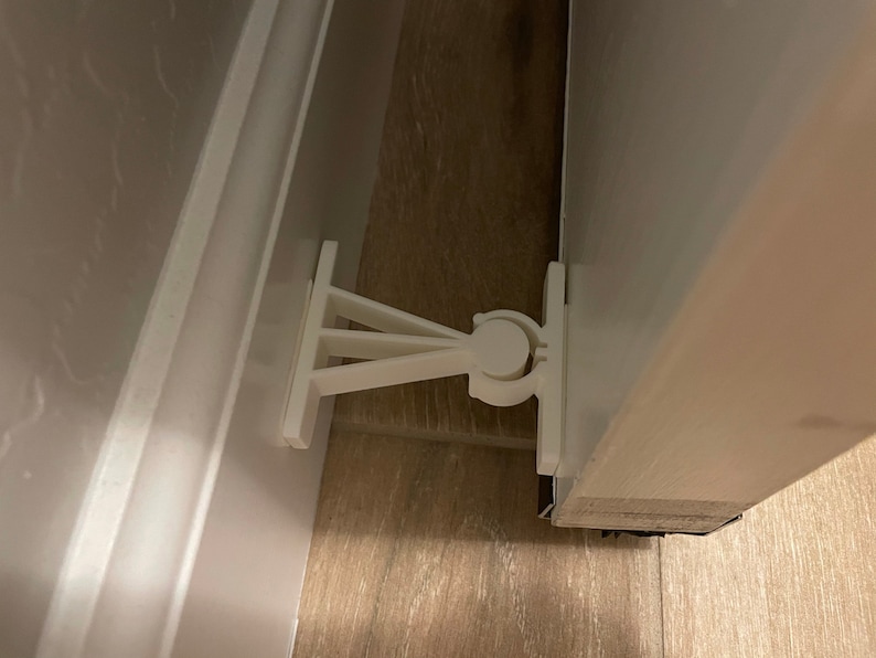 May include: White plastic door stop with a rounded knob and a triangular base. The door stop is attached to a white door frame and a white baseboard.