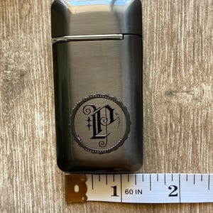 Puede incluir: Un encendedor de metal gris oscuro con un emblema decorativo con la letra "P" en un dise&ntilde;o circular. El encendedor mide aproximadamente 5 cm de largo. El encendedor est&aacute; sobre una superficie de madera.