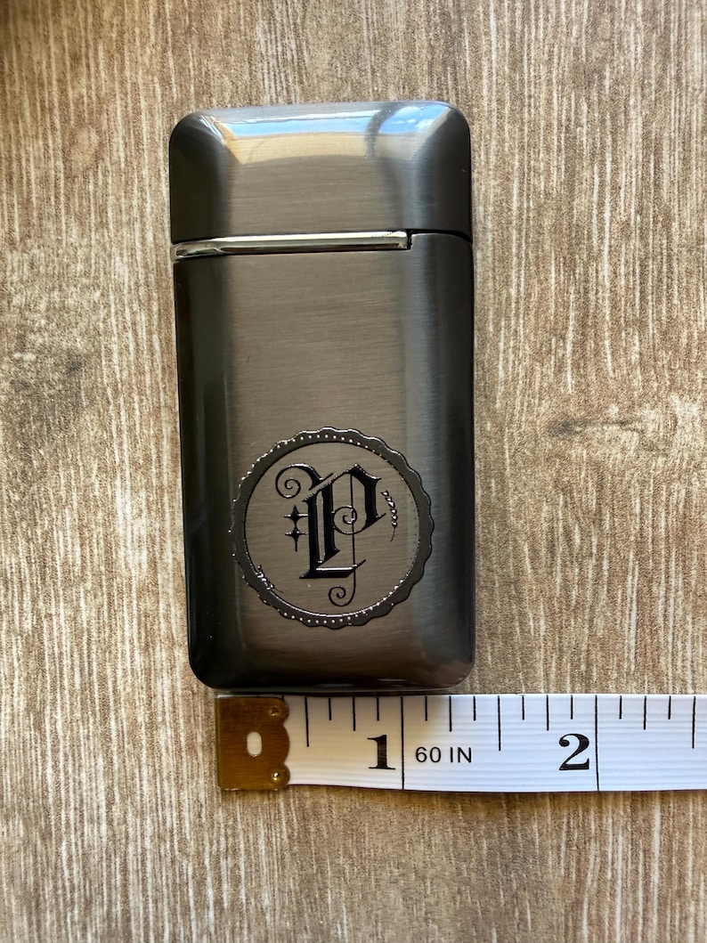 Puede incluir: Un encendedor de metal gris oscuro con un emblema decorativo con la letra "P" en un dise&ntilde;o circular. El encendedor mide aproximadamente 5 cm de largo. El encendedor est&aacute; sobre una superficie de madera.