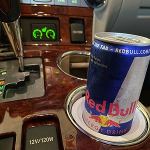 May include: A silver can of Red Bull energy drink with a blue and white label is in a cup holder in a car. The can is cold and has condensation on it. The text on the can reads "THE TAB - REDBULL.COM/SO" and "ENERGY DRINK".