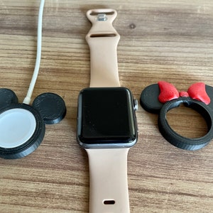 May include: An Apple Watch with a tan band rests on a wooden surface, next to a black charging stand with a white charging disc and a Minnie Mouse-themed watch stand with a red bow. The watch face is black.