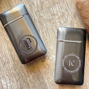 Puede incluir: Dos pitilleras de metal gris plomo con detalles plateados. Una pitillera tiene un monograma "P" decorativo, y la otra "JC" en un dise&ntilde;o circular blanco. Las pitilleras est&aacute;n sobre una superficie de madera.