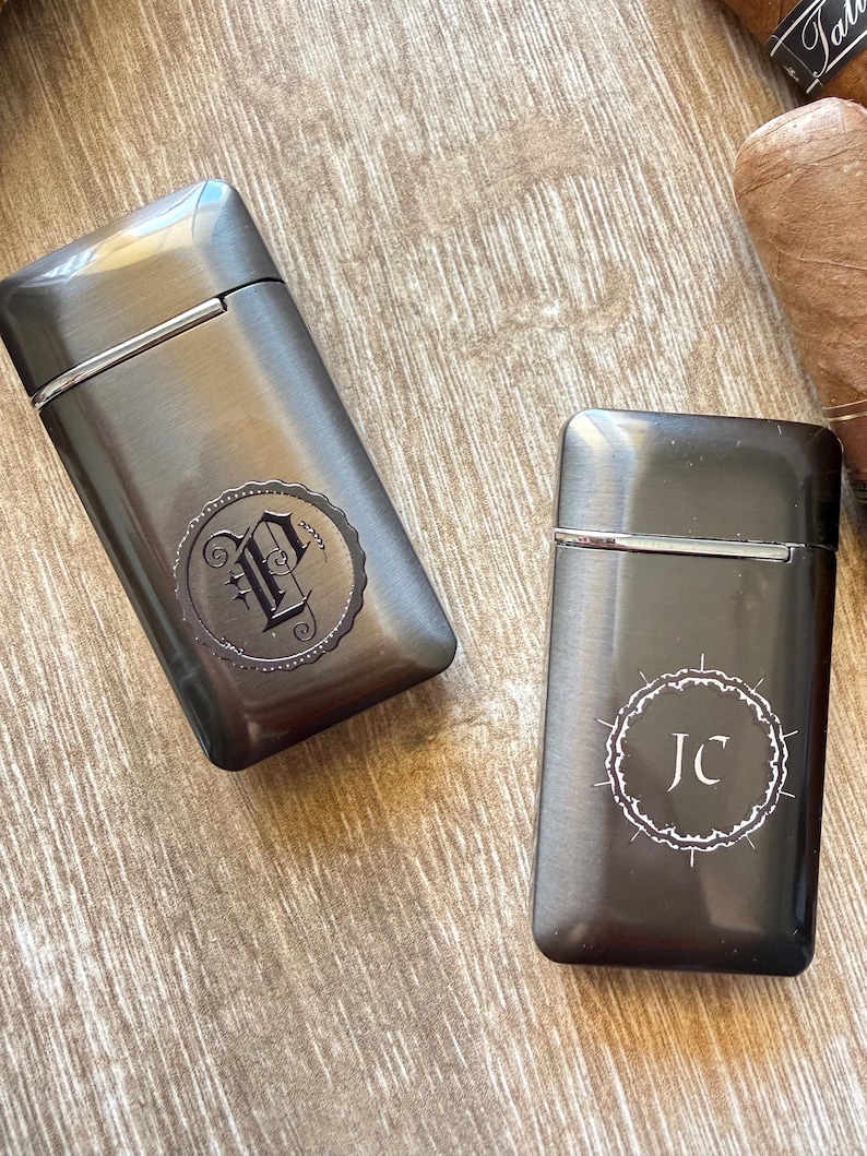 Puede incluir: Dos pitilleras de metal gris plomo con detalles plateados. Una pitillera tiene un monograma "P" decorativo, y la otra "JC" en un dise&ntilde;o circular blanco. Las pitilleras est&aacute;n sobre una superficie de madera.