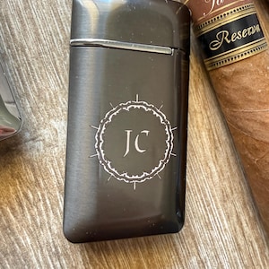 Puede incluir: Un encendedor de metal gris oscuro con las iniciales "JC" dentro de un c&iacute;rculo decorativo blanco. El encendedor es rectangular con un borde plateado. Un cigarro con una etiqueta marr&oacute;n y negra est&aacute; en el fondo.