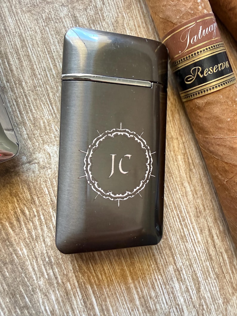 Puede incluir: Un encendedor de metal gris oscuro con las iniciales "JC" dentro de un c&iacute;rculo decorativo blanco. El encendedor es rectangular con un borde plateado. Un cigarro con una etiqueta marr&oacute;n y negra est&aacute; en el fondo.