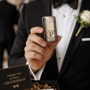 Puede incluir: Un encendedor de metal plateado con una inicial "P" grabada es sostenido por una persona que lleva un esmoquin negro con pajarita. Una tarjeta que dice "THANK YOU for being my GROOMSMAN" es visible en una caja de regalo. Un ojal de rosa blanca est&aacute; prendido al esmoquin.