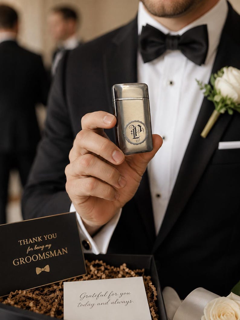 Puede incluir: Un encendedor de metal plateado con una inicial "P" grabada es sostenido por una persona que lleva un esmoquin negro con pajarita. Una tarjeta que dice "THANK YOU for being my GROOMSMAN" es visible en una caja de regalo. Un ojal de rosa blanca est&aacute; prendido al esmoquin.
