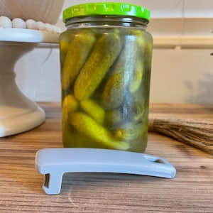 Peut inclure: Un bocal en verre rempli de cornichons à l'aneth, avec un couvercle vert et un ouvre-bocal en plastique gris.