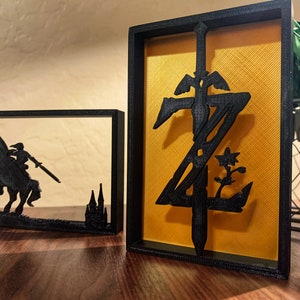 May include: Two black framed shadow boxes with a gold background. The left box features a silhouette of a knight riding a horse with a sword. The right box features a stylized sword with wings and a flower.