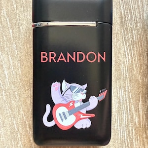 Puede incluir: Encendedor negro con el nombre "BRANDON" en letras rojas. Un gato de dibujos animados con gafas de sol y tocando una guitarra el&eacute;ctrica roja aparece debajo del nombre. El encendedor tiene un borde plateado.