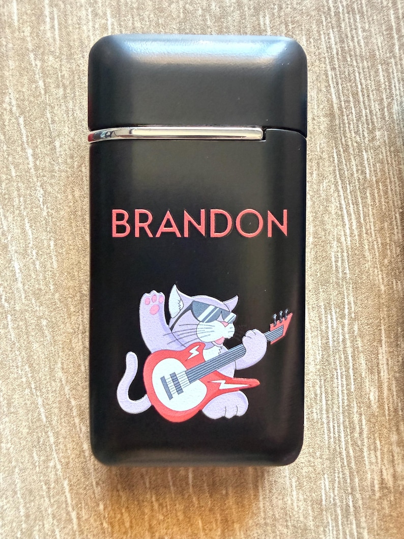 Puede incluir: Encendedor negro con el nombre "BRANDON" en letras rojas. Un gato de dibujos animados con gafas de sol y tocando una guitarra el&eacute;ctrica roja aparece debajo del nombre. El encendedor tiene un borde plateado.