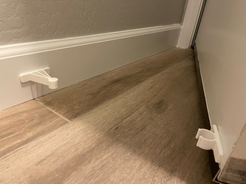 May include: White plastic door stops installed on a wood floor. One door stop is mounted to the wall and the other is mounted to the door.