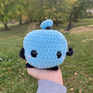 May include: A light blue crocheted plush toy with black accents, including small arms, feet, and a top. The toy has large black eyes and pink cheeks, giving it a cute appearance. The plush is held in a person's hand against a blurred green background.