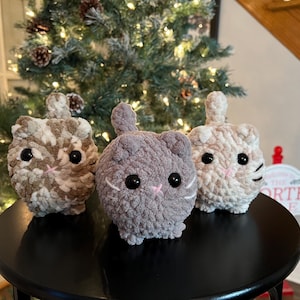 May include: Three plush cat toys in shades of gray, beige, and cream sit on a black surface. Each cat has large black eyes, a pink nose, and a small tail. The background includes a Christmas tree.