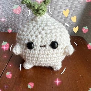 May include: A white crocheted plush toy shaped like a turnip with a green crocheted stem and black eyes. The toy is sitting on a wooden surface with a strawberry pattern.