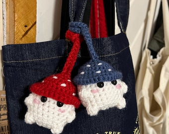 May include: Two crocheted mushroom plushies with white bodies, red and blue hats, and black eyes. The plushies are attached to a blue denim tote bag with the text "And if that's true... Sabrina" printed on the bag.